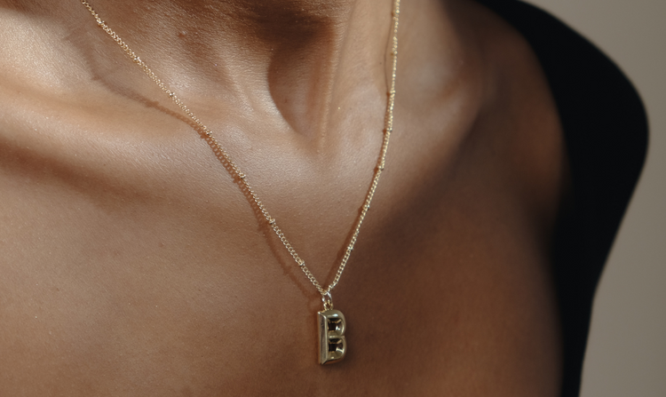 A person wears a delicate gold necklace with a bold letter "B" pendant.