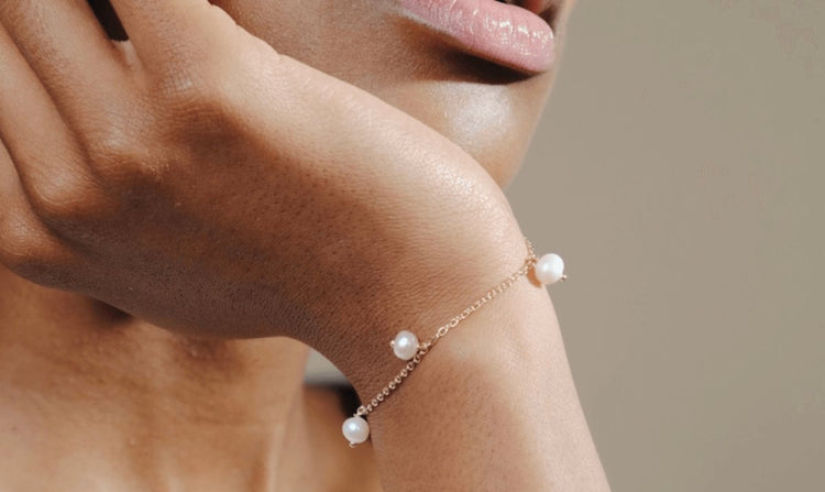 A woman rests her face on her hand, showcasing a delicate gold bracelet adorned with small pearls.