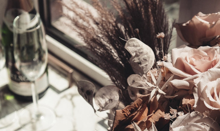 A still-life photograph features champagne glasses and a bottle beside a floral arrangement of roses and dried grasses near a window.