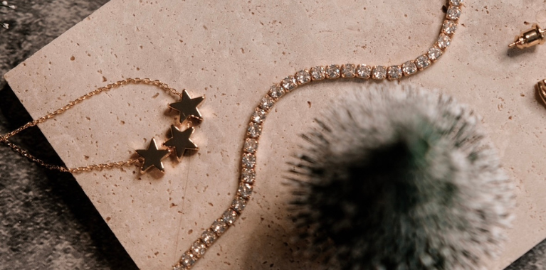 Close-up of gold-toned jewelry, including a star necklace and a tennis bracelet, on a textured surface with a blurred decorative object.