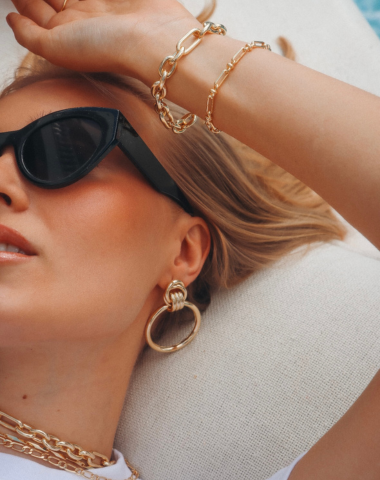 Close-up of a woman wearing sunglasses and gold jewelry, including bracelets, earrings, and a necklace.