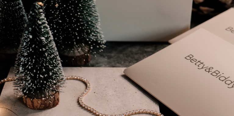 Three small, snow-flocked Christmas trees sit beside a gold-colored necklace and cards with the text