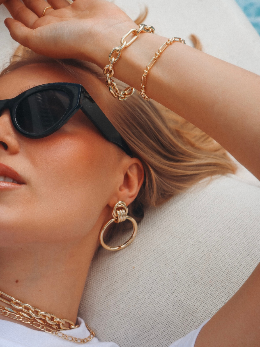 Close up of a blonde woman wearing sunglasses and gold jewelry, including a Helix Bracelet, bracelets, earrings, and a necklace.