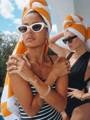 Two women wearing swimsuits, jewelry, sunglasses, and orange-and-white striped towels on their heads, also wearing Alexa Gold Hoops.