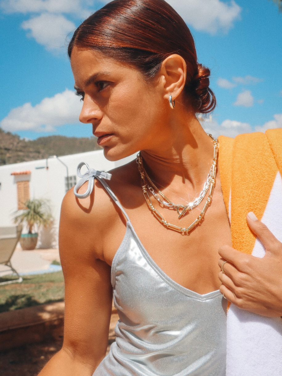 A woman with auburn hair wearing an Inter Curb Toggle T-Bar and layered necklaces holds a towel against a bright blue sky.