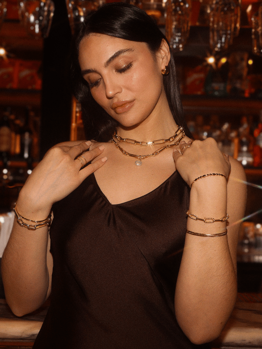 A woman in a dark brown slip dress poses while wearing layered gold necklaces and bracelets, including an Aura Gold Bangle.