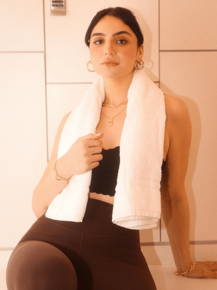 Woman with towel draped over her shoulders, wearing workout attire and jewelry, including the Omni Evil Eye Coin, sitting against a tiled wall.