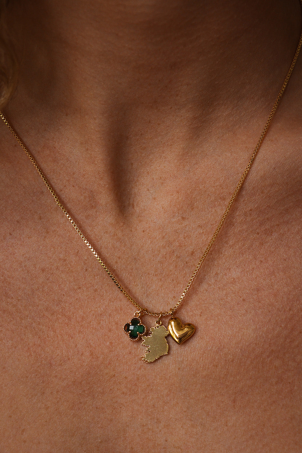 Close-up of a woman's chest and the gold Eire Charm Necklace she is wearing with three charms: a green clover, a map of Ireland, and a heart.