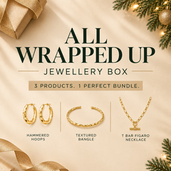 'All Wrapped Up Jewellery Box' featuring three gold jewelry items: hammered hoops, a textured bangle, and a T-bar figaro necklace, arranged on a beige background with gold ribbon, gift box corner, and pine branches.