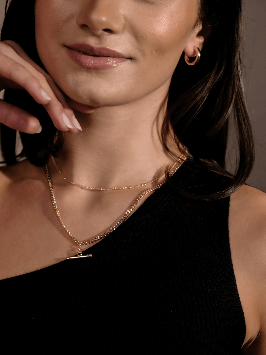 A woman wearing a gold hoop earring and two gold necklaces, including a T-Bar Curb Chain, poses with her hand near her face.