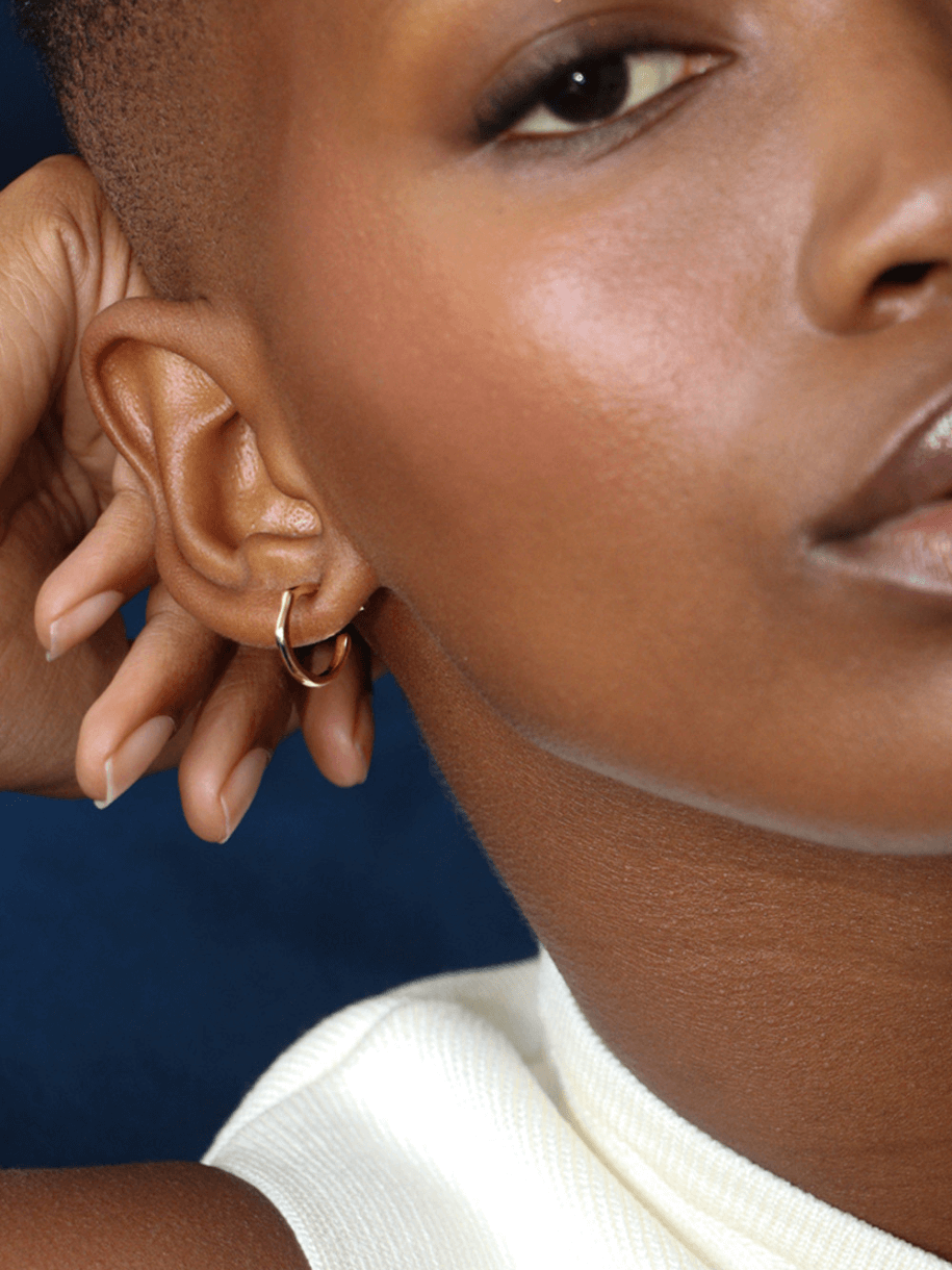 Close-up of a dark-skinned person wearing a small gold The Dented Huggie Hoop earring.