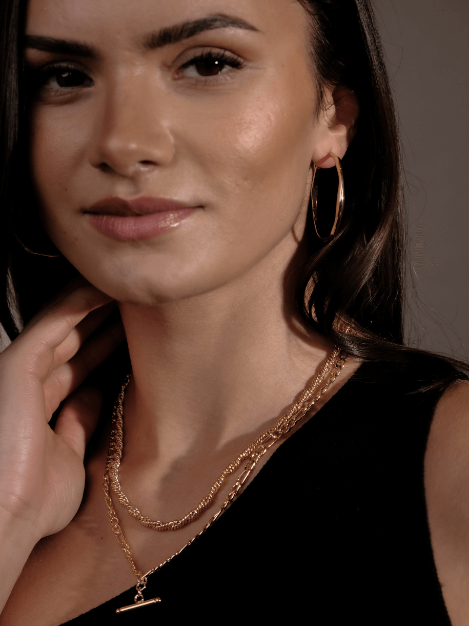 Close-up of a woman wearing gold hoop earrings and a layered gold T-Bar Figaro Necklace.