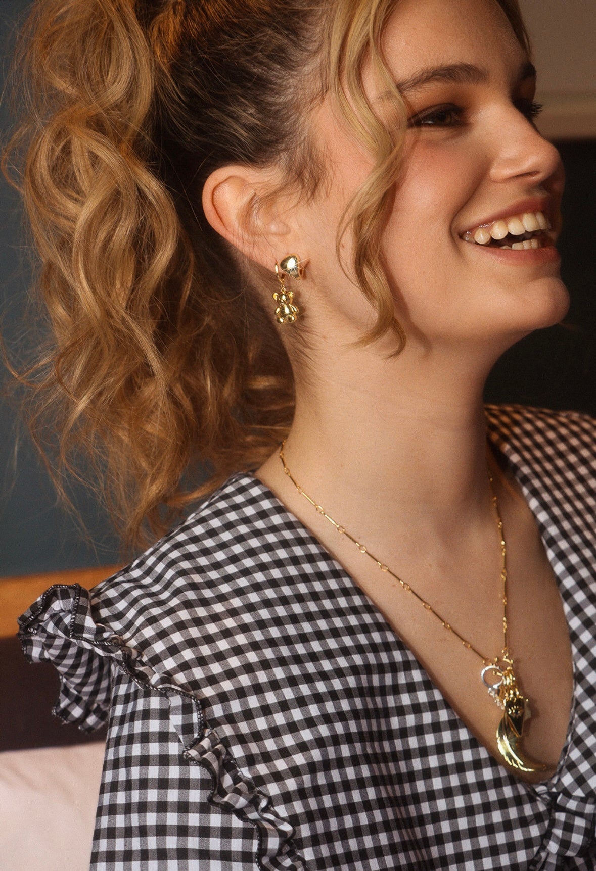 A smiling woman with blonde curly hair, wearing gold jewelry and a black-and-white gingham dress.