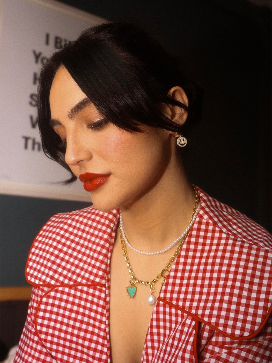A woman in a red and white checkered shirt wearing Smiley Face Charm jewelry looks downward.