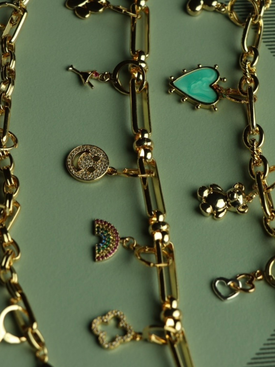 Close-up of gold Smiley Face Charm bracelets with various pendant shapes on a green surface.