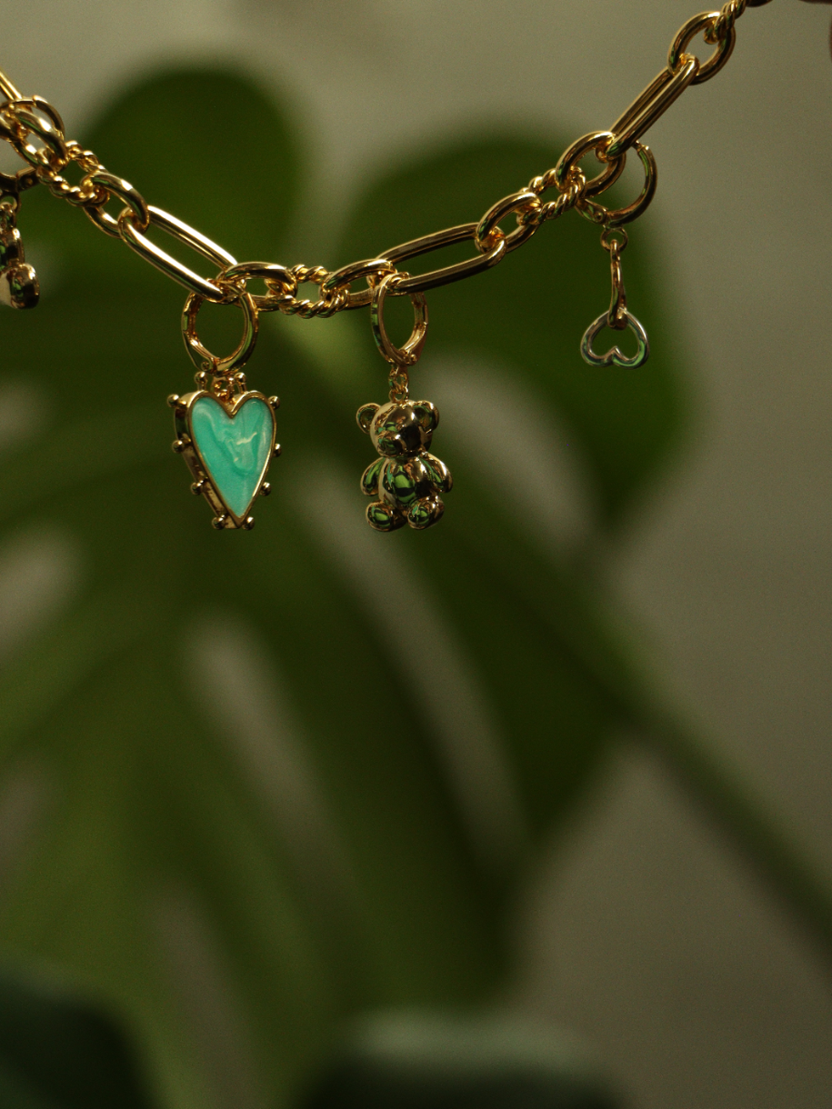 A gold Teddy Bear Charm chain with assorted charms dangles in front of green foliage.