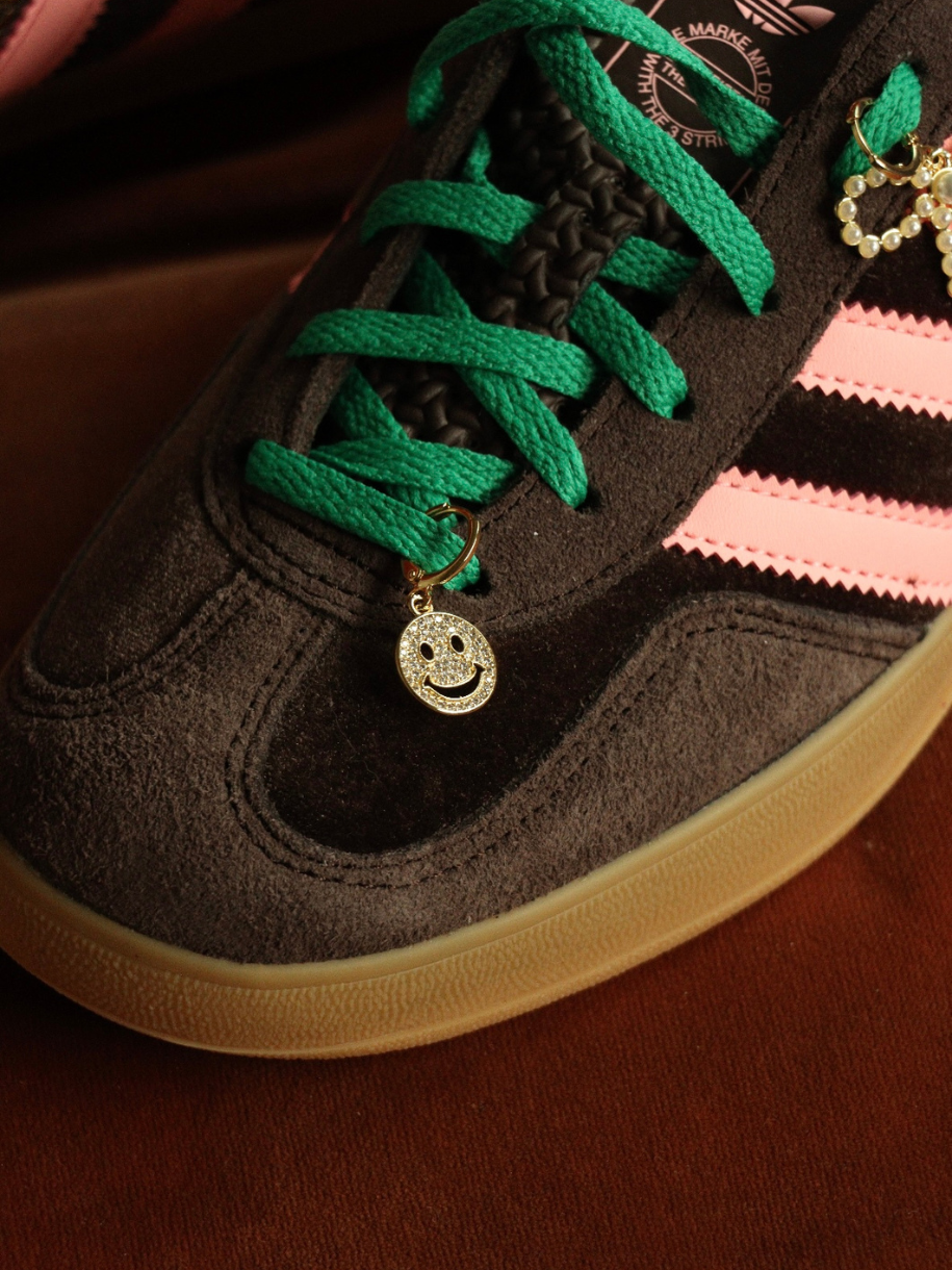 Close-up of a brown suede Adidas sneaker with pink stripes, green and black laces, and Smiley Face Charm and bow charms.
