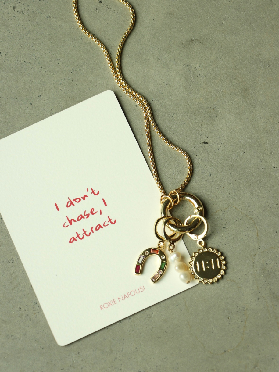 A gold 11:11 Charm Pendant Gold necklace with a horseshoe, pearls, and a sunburst with "11:11" hangs beside a card that reads "I don't chase, I attract, ROXIE NAFOUSI."