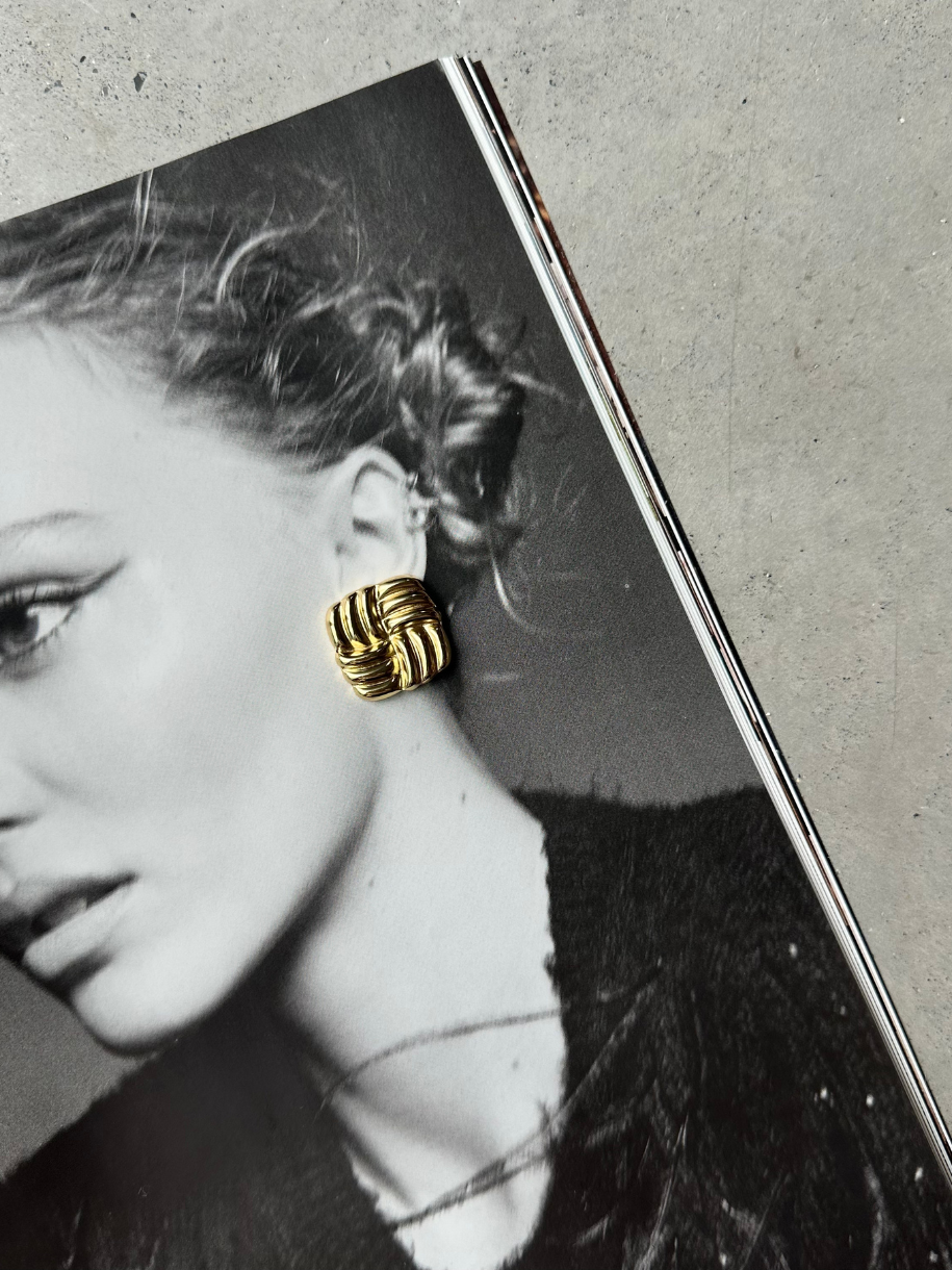 A gold, square, woven-patterned Sable Stud earring rests on a black-and-white magazine page featuring a woman's portrait.