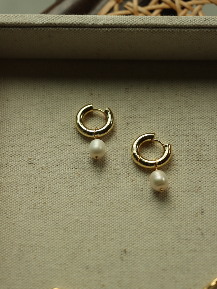 A pair of gold The Chunky Pearl Hoop earrings with pearl drops rest on a tan textured surface.