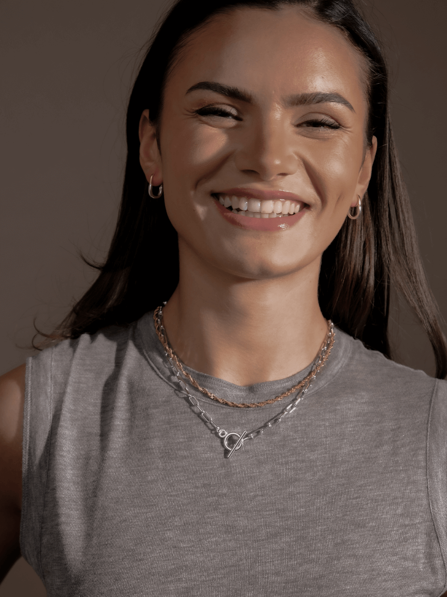 A smiling woman with dark hair wearing two Chunky Link and Round Hook Toggle Silver necklaces and gold hoop earrings.