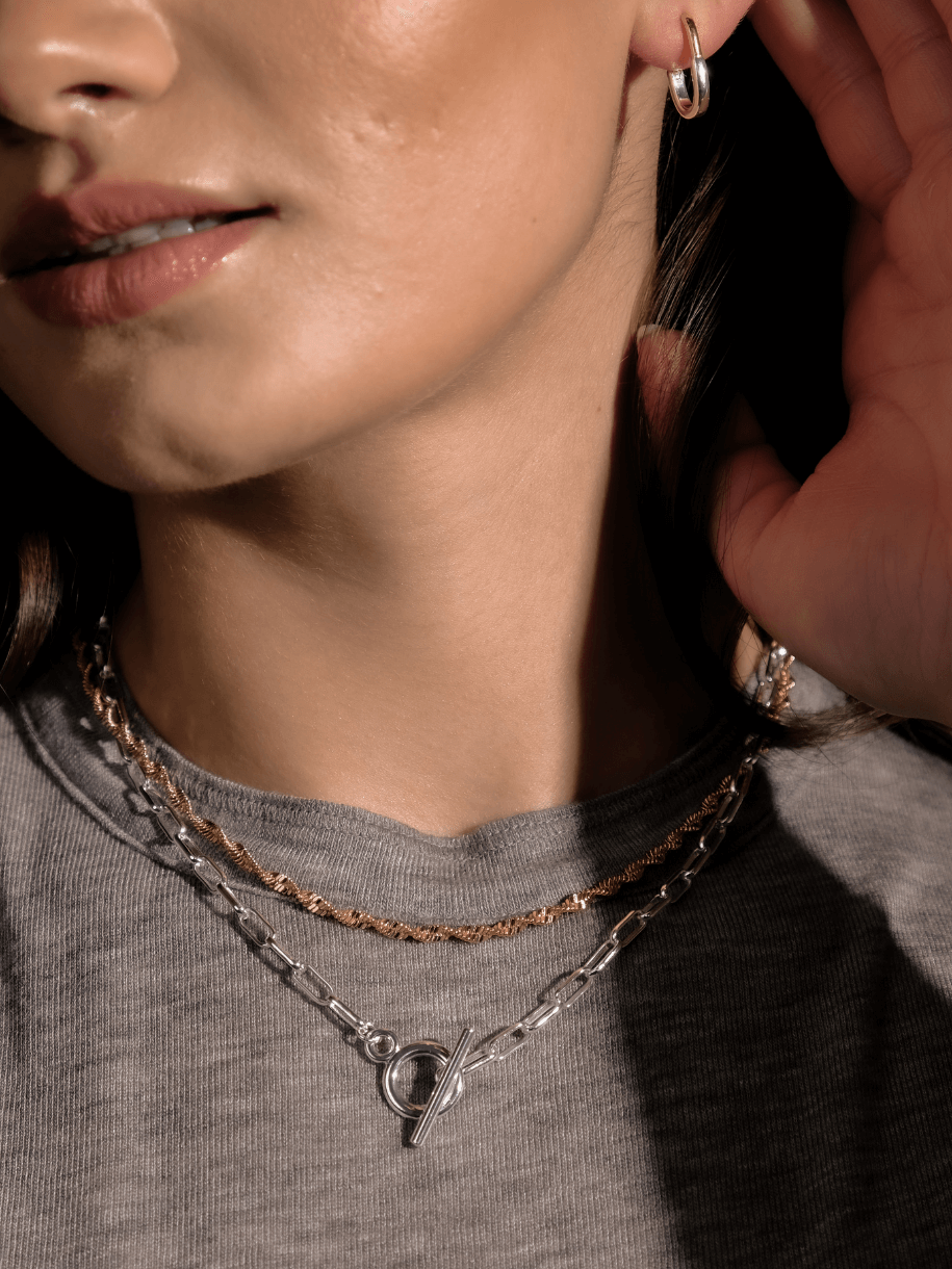 A person wearing Chunky Link and Round Hook Toggle Silver hoop earrings and two necklaces against a gray shirt.