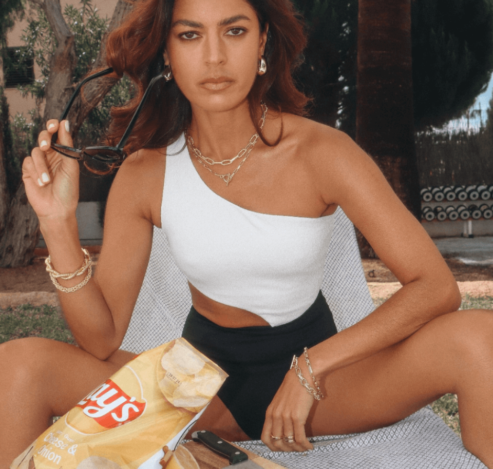 A woman in a one-shoulder swimsuit poses with a bag of chips and jewelry outdoors.