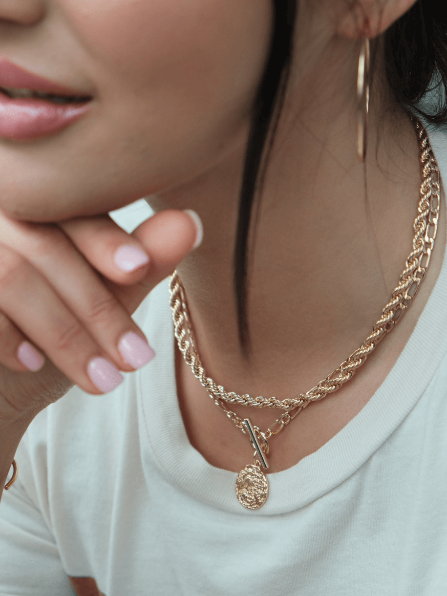 A woman wearing a white shirt, gold hoop earrings, and two gold necklaces, including the Athena Toggle Coin, with her hand near her chin.