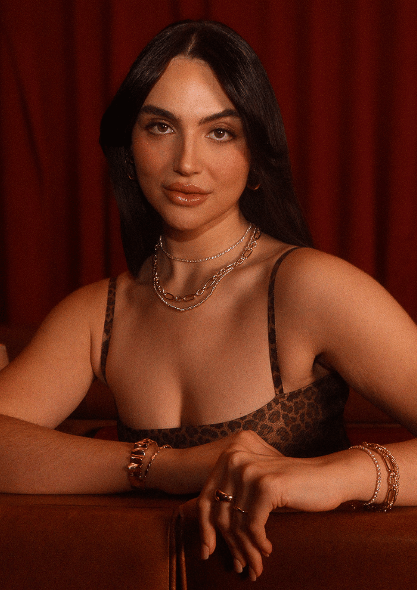 A woman with long dark hair and gold jewelry wearing a Calver Block Bangle sits in front of a red backdrop.