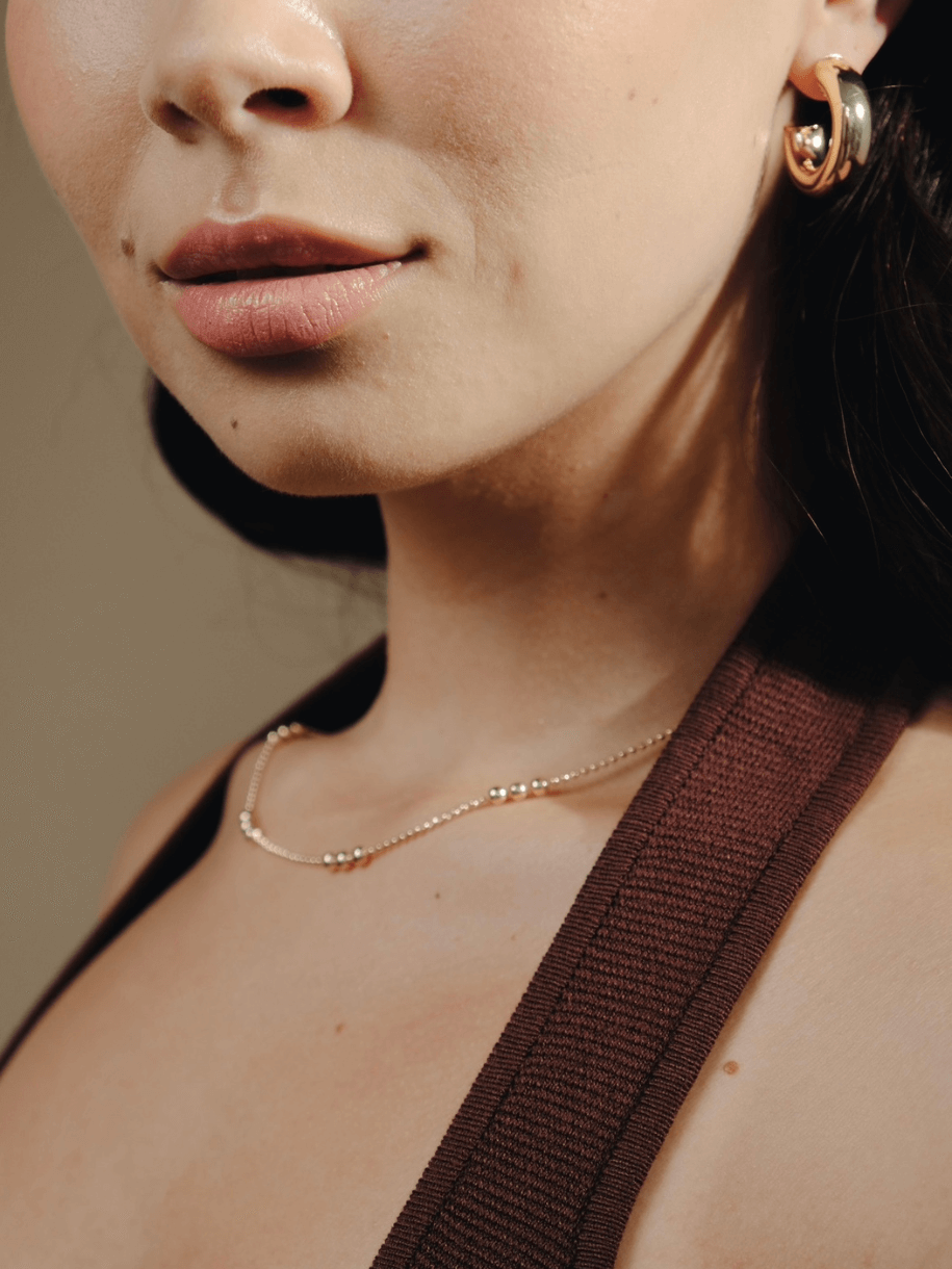 Close-up of a woman wearing The Dotted Ball Necklace 14 inches and gold earrings, with brown clothing straps visible.