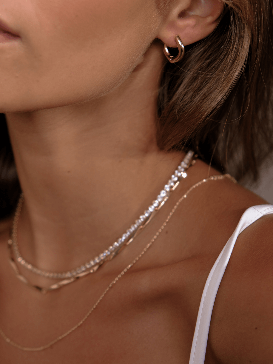 Close-up of a woman's neck and shoulder wearing layered necklaces and a hoop earring, including a Tennis Necklace - Clear Gemstone.