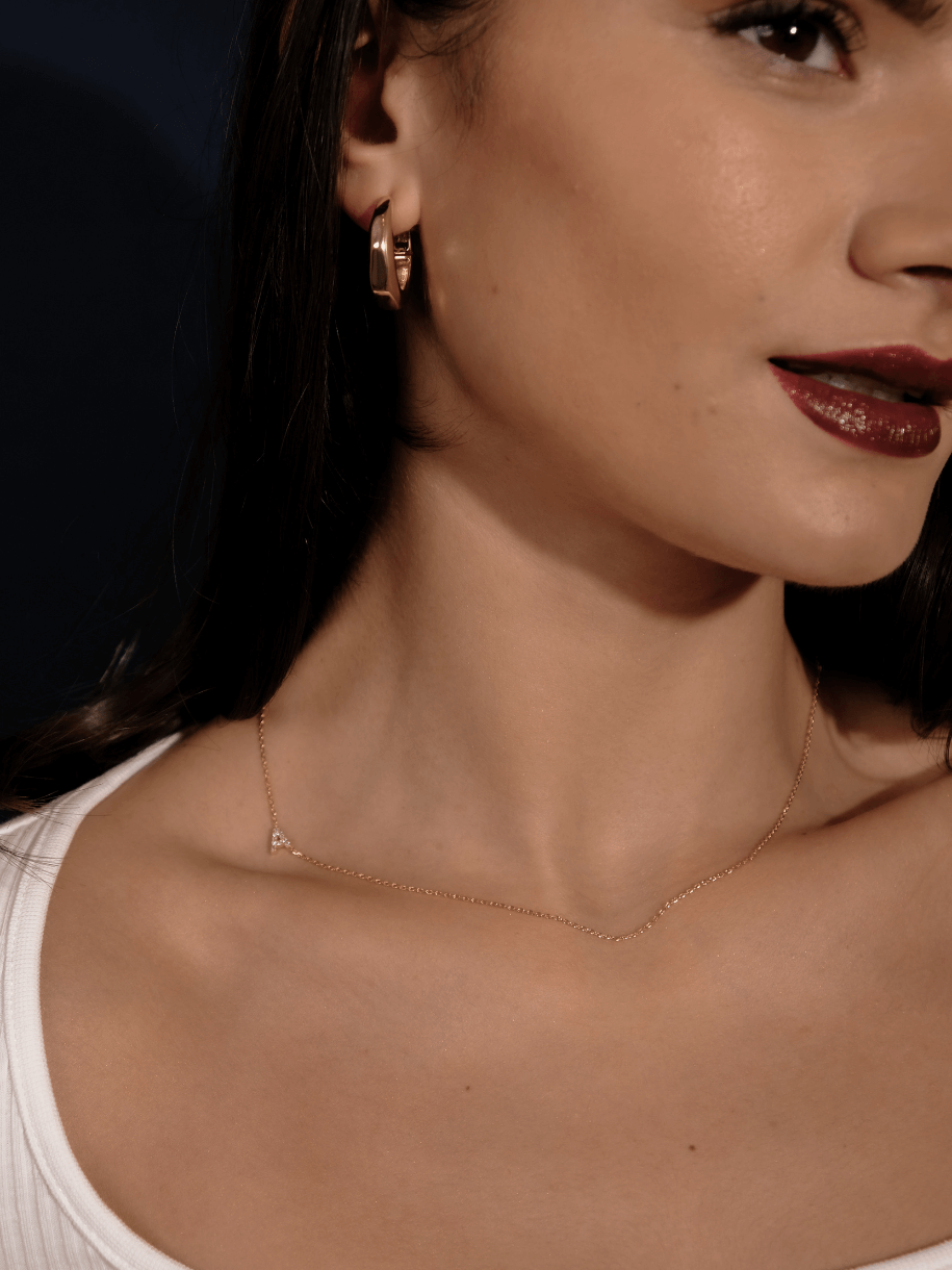 Close-up of a woman with dark hair, wearing The Diamante Initials gold jewelry, including earrings and a necklace, and dark red lipstick.