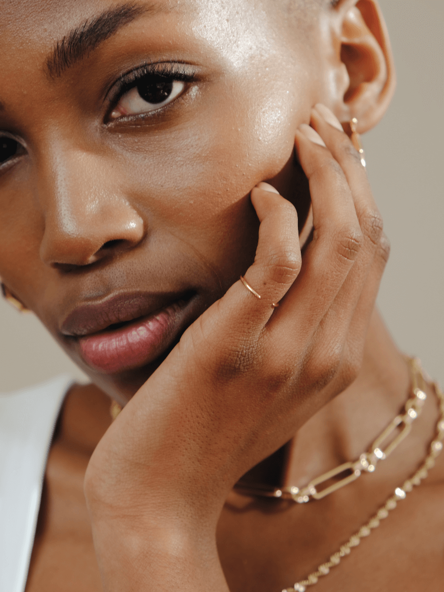 Close-up of a Black woman with short hair, wearing gold jewelry and holding her face.