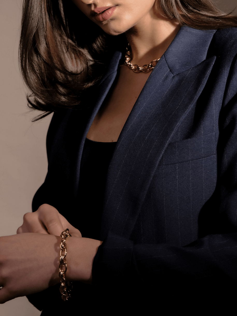 A person in a pinstripe blazer adjusts a gold chain Oval Cross Link Bracelet, wearing a matching necklace.