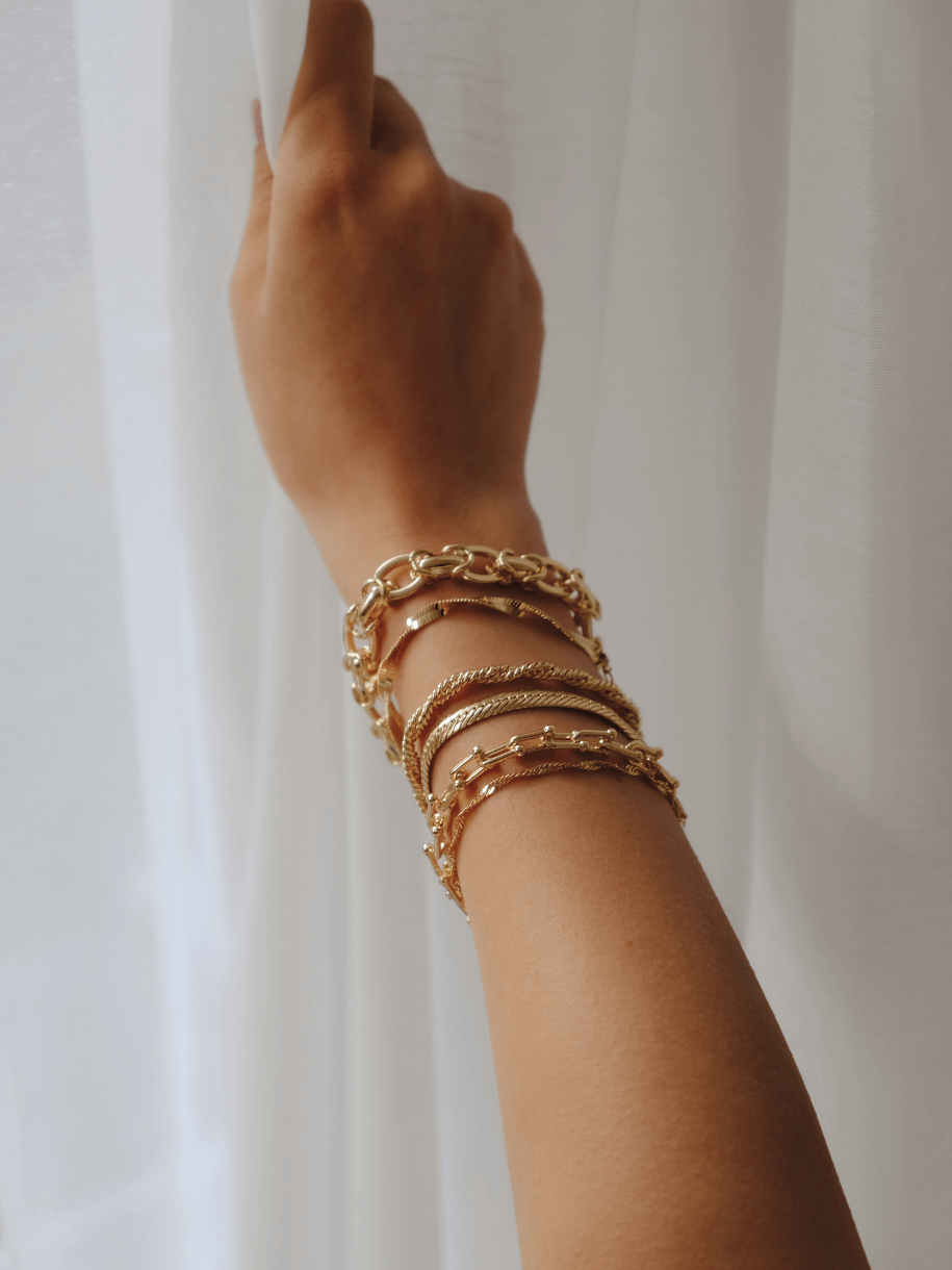 Arm with multiple gold Snake Herringbone Bracelets against a white curtain backdrop.