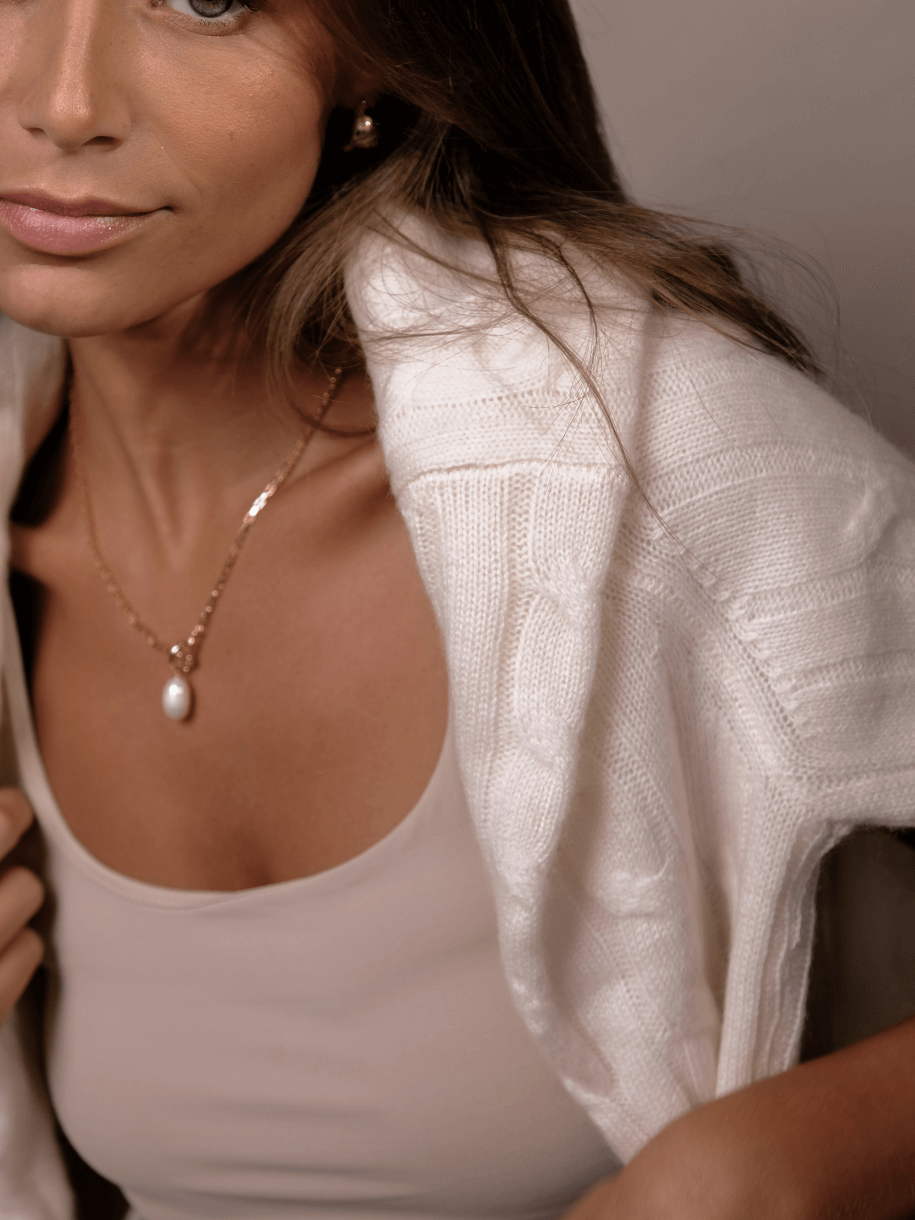 Close-up of a woman wearing a gold necklace with a pearl pendant and a white cable knit sweater.