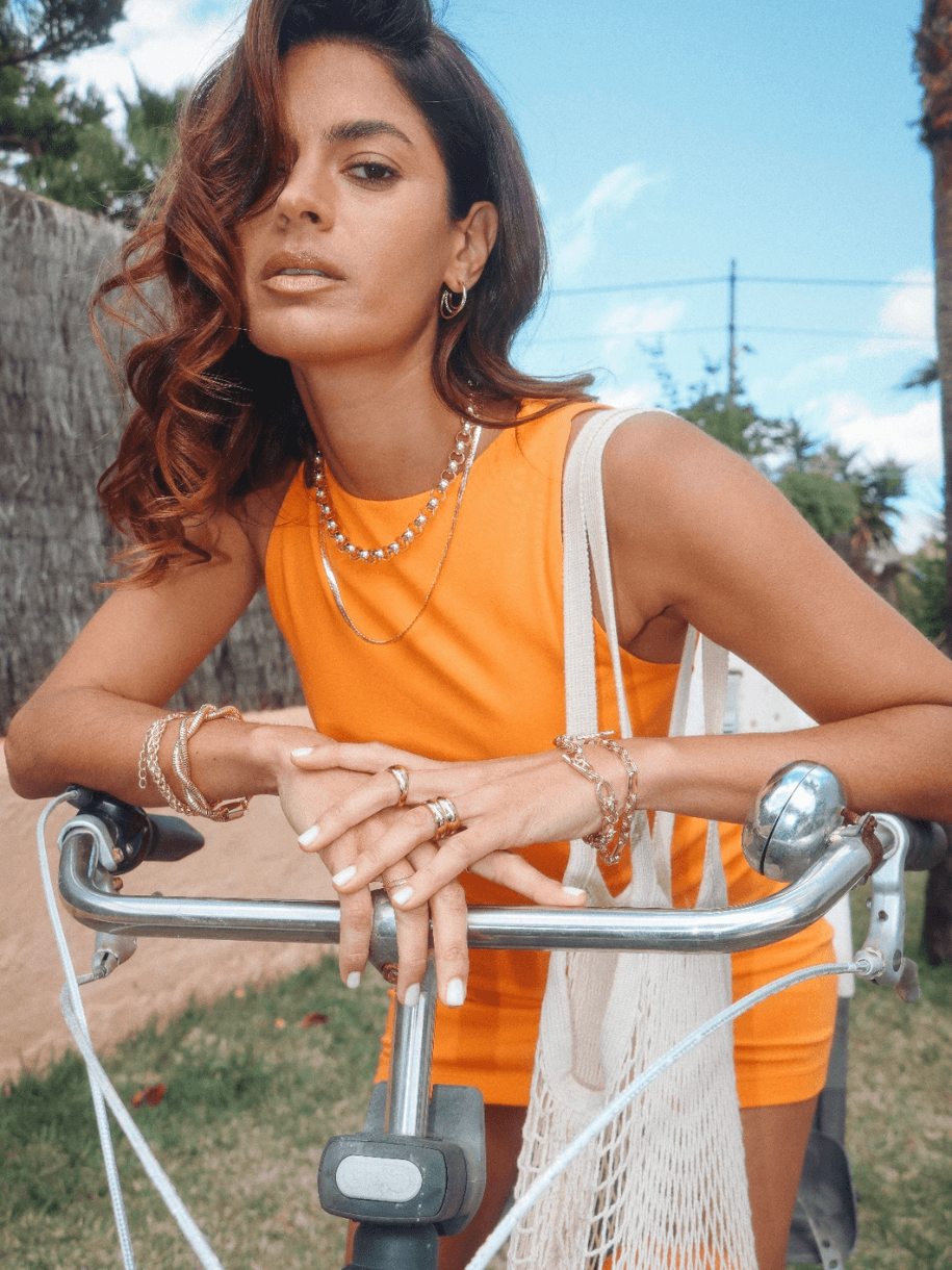 A woman in an orange dress and jewelry poses with her hands on the handlebars of a bicycle, wearing a Rolo Necklace.