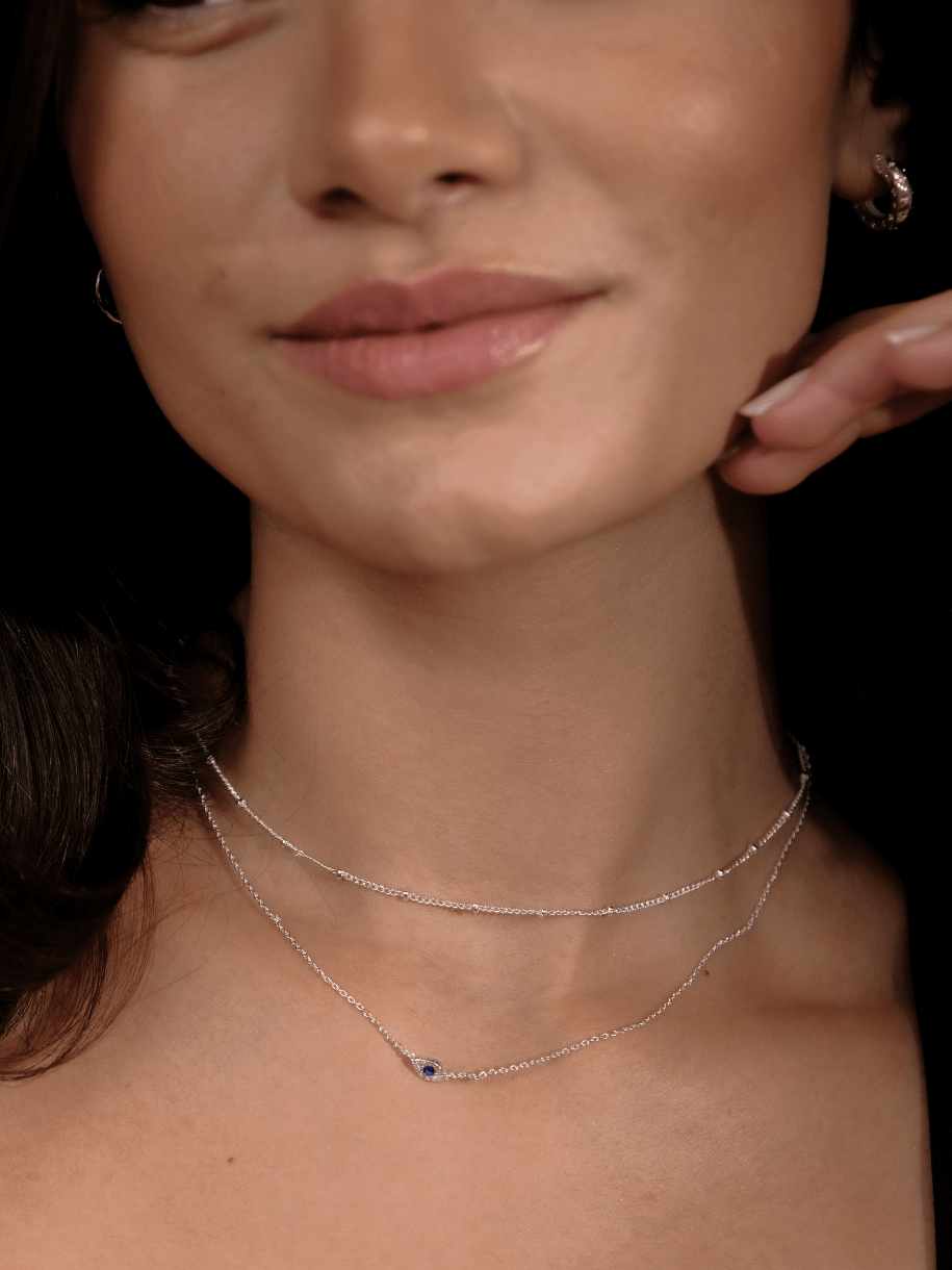 A woman's face and neck are shown wearing two delicate silver necklaces, one with a blue pendant, including the Single Beaded Chain 14- Silver.