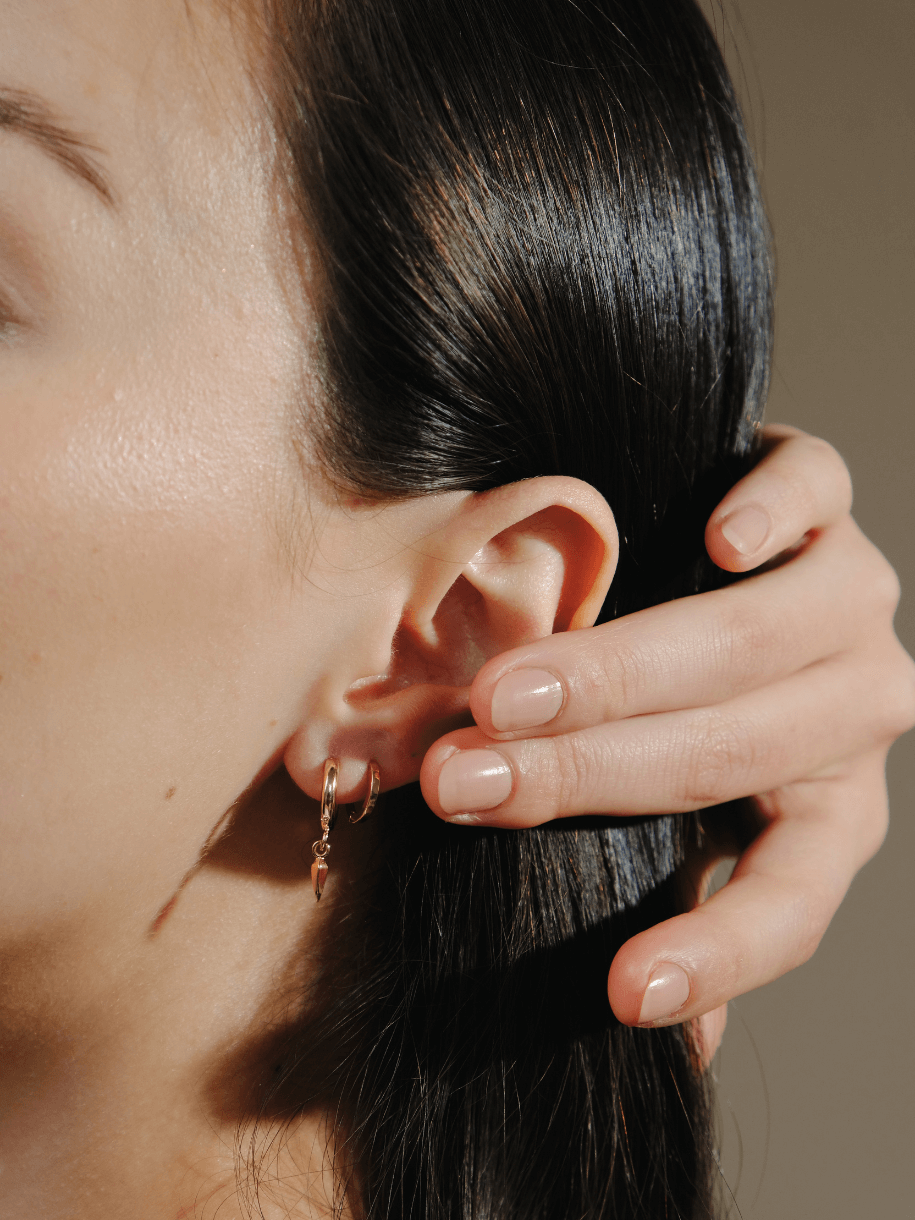 Close-up of a woman's ear with gold Drop Claw Huggie earrings and her hand touching her dark hair.