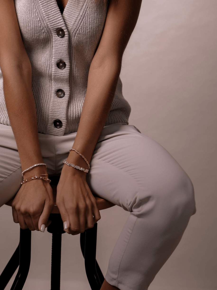 A person wearing a Crossing Twist Bracelet, a knit vest, and pants sits on a stool.