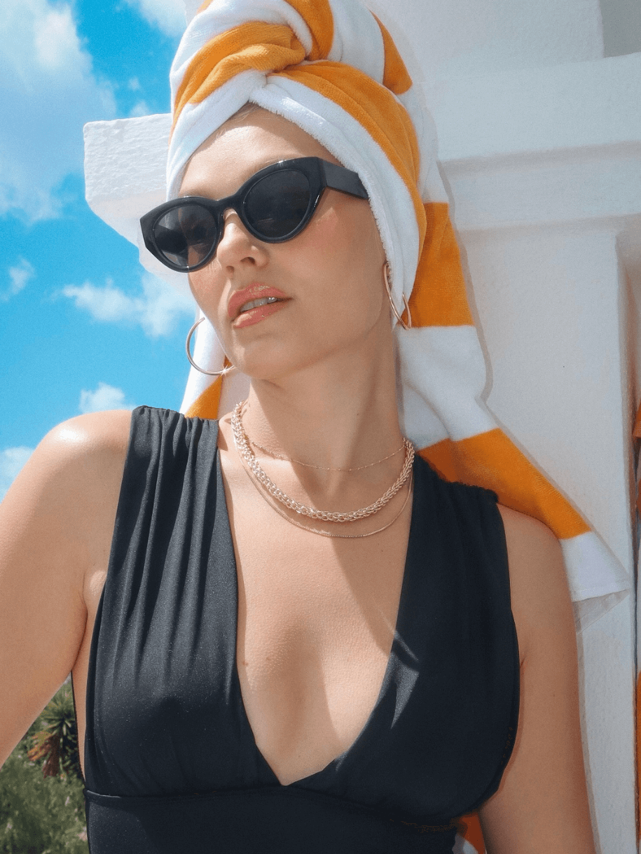 A woman in a black bathing suit, sunglasses, and towel wrapped on her head poses against a blue sky, possibly wearing the Thin Beaded Chain 14 inches.