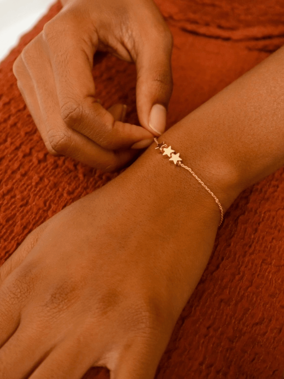 A hand fastens a gold Trio Star Charm Bracelet.