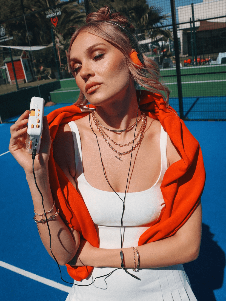 Woman in a white tennis outfit with a bright orange sweater around her shoulders, listening to a portable cassette player on a blue surface, with a Block Wave Bangle.