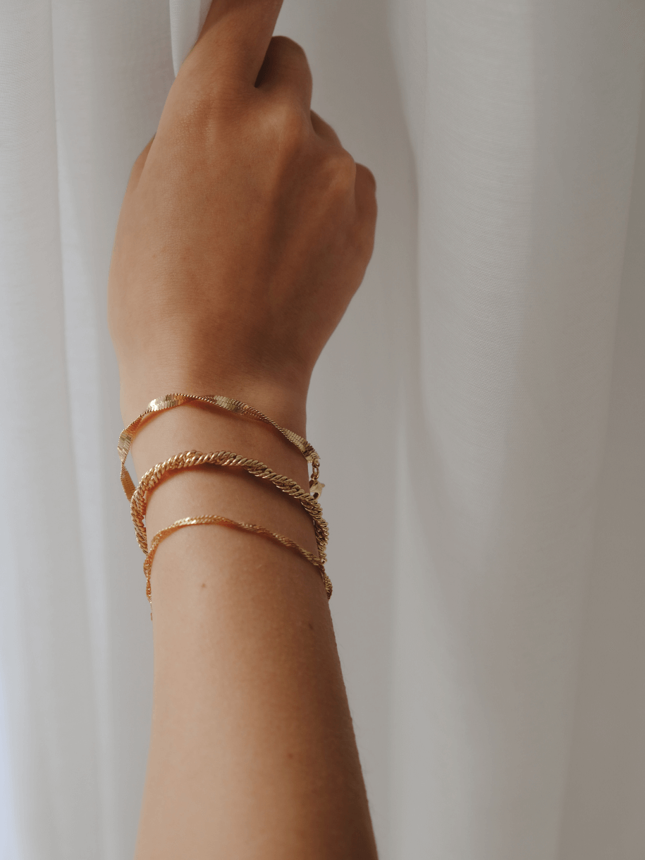 Arm wearing three gold bracelets, including The Twisted Chain Bracelet, against a white curtain.