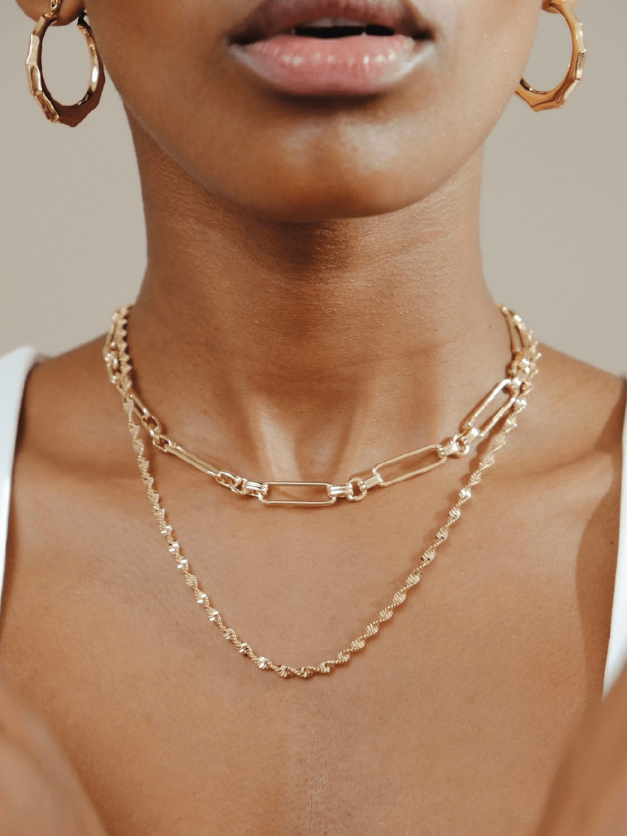 Close-up of a person wearing a Twisted Herringbone Necklace, two gold necklaces, and gold hoop earrings.