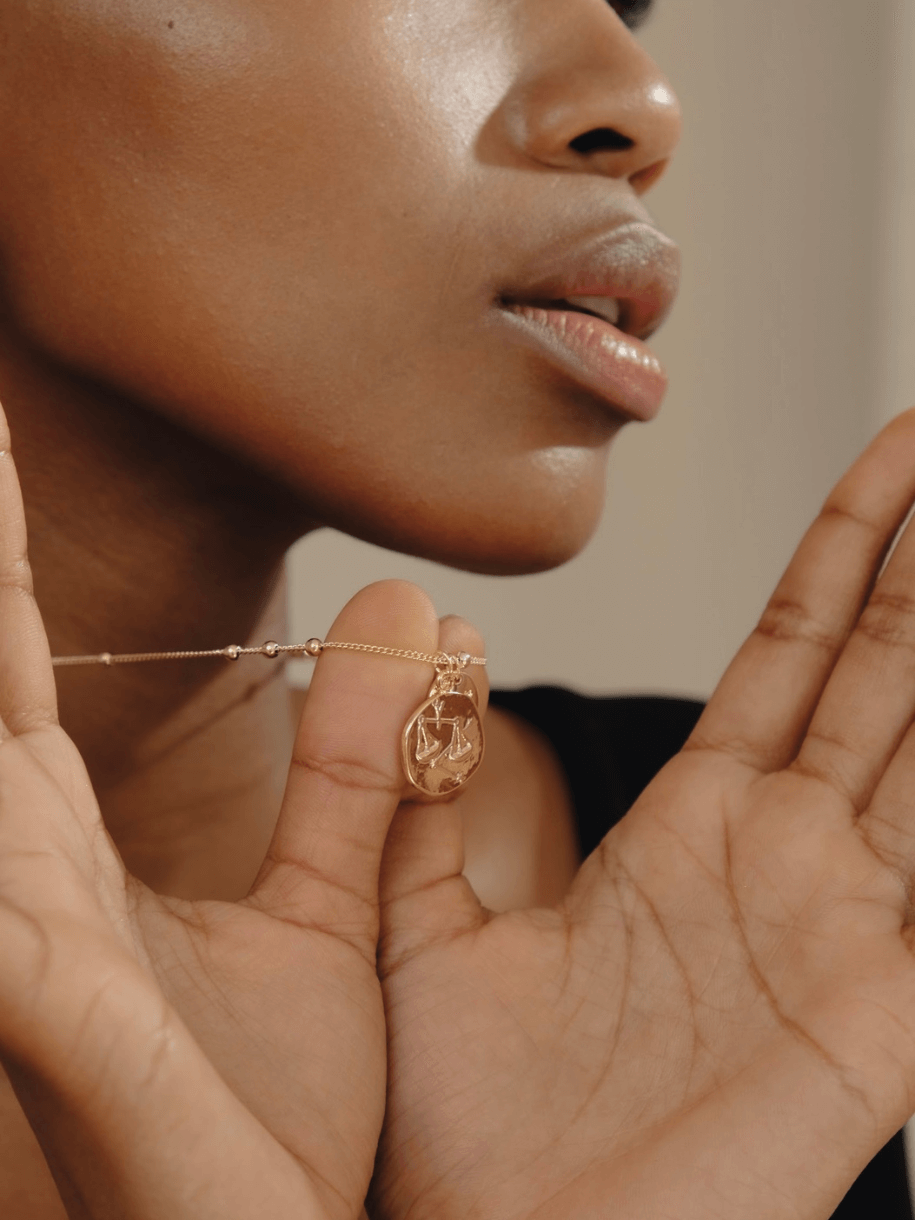 A person with dark skin holding a gold necklace with a Scorpio Zodiac Coin pendant.
