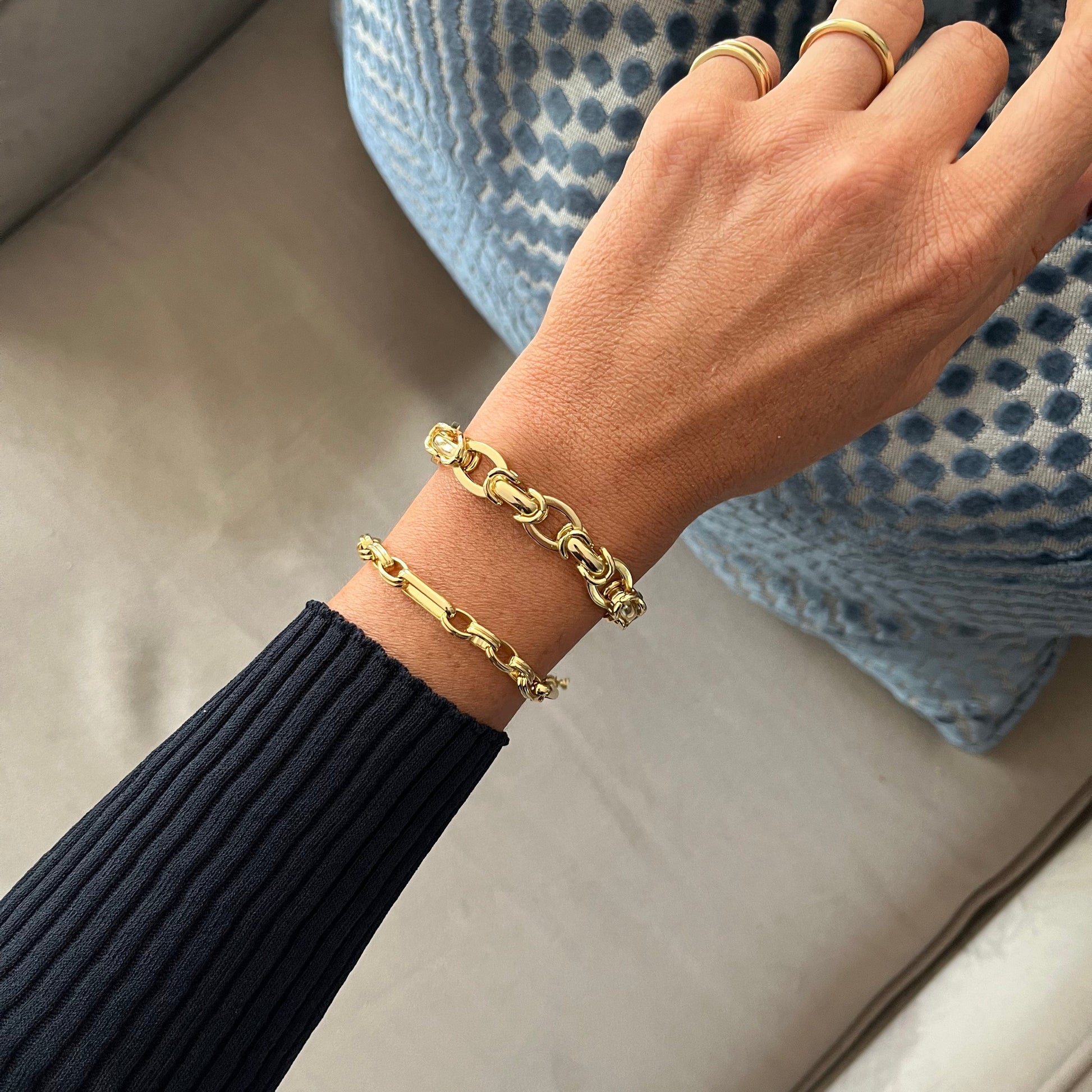A person's arm rests on a couch, adorned with a Triple Link Bracelet, two gold bracelets and rings.