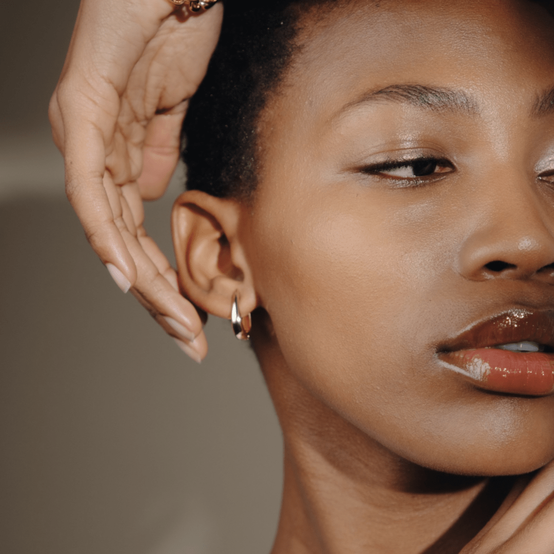 Close-up of a person with a Claw Hoop earring and shiny lips, with a hand near their face.