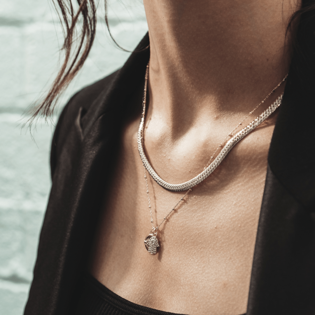 A person wearing two layered necklaces, one a The Snake Herringbone 14 inches style and the other with a small charm, with a black jacket.