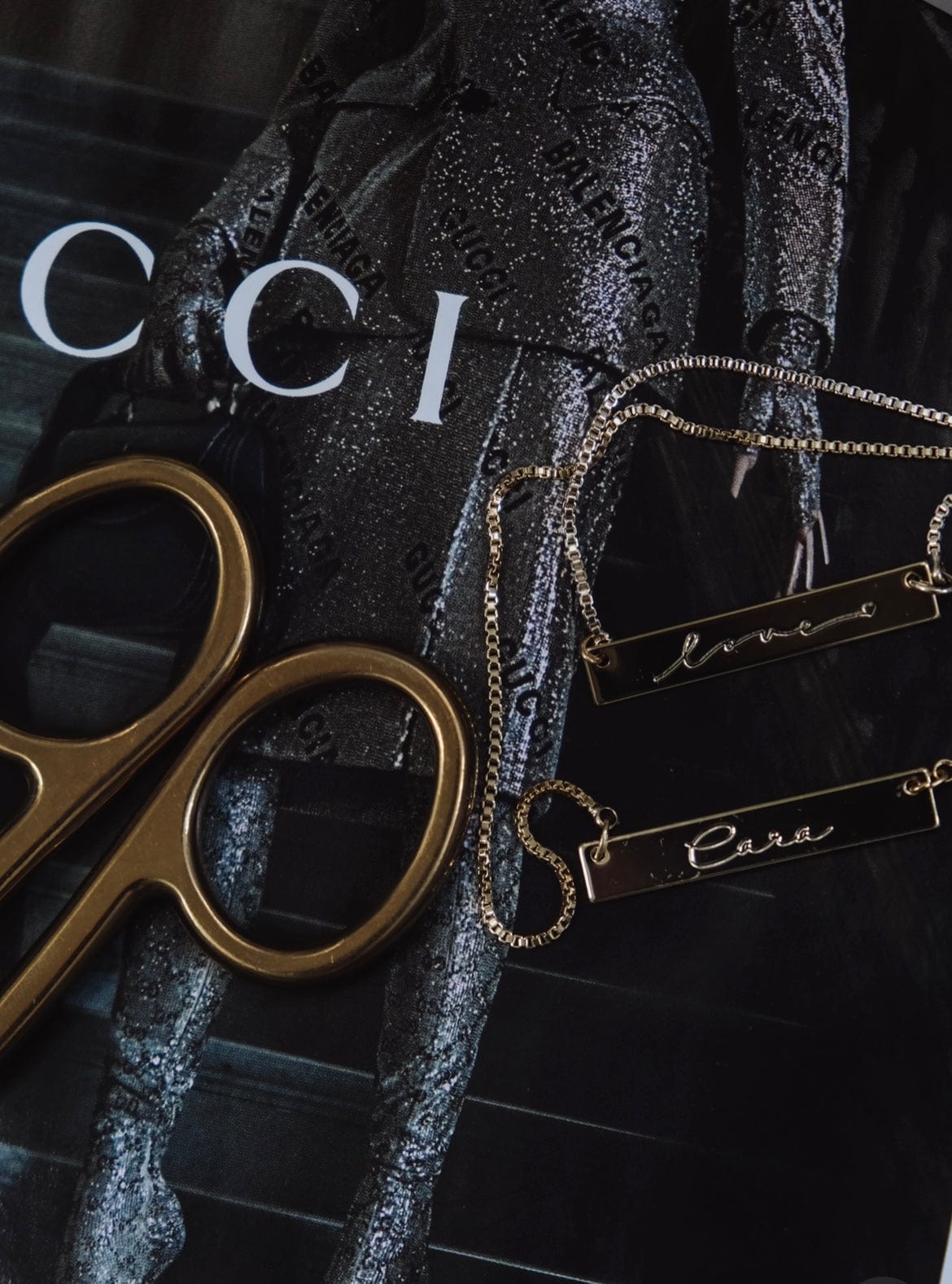 Gold scissors and two gold necklaces rest on a magazine featuring a figure in a sparkly outfit with brand names near a Cara Bar Necklace.